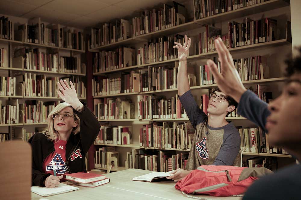 A group of students sit at a table in a library with their hands raised.
