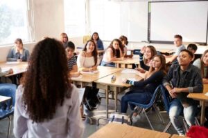 A diverse group of high school students sit at desks in a classroom.