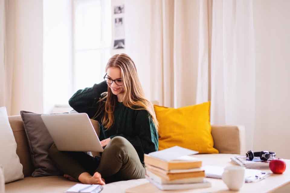 A smiling college student surrounded by books and notebooks sits on a sofa using a laptop.