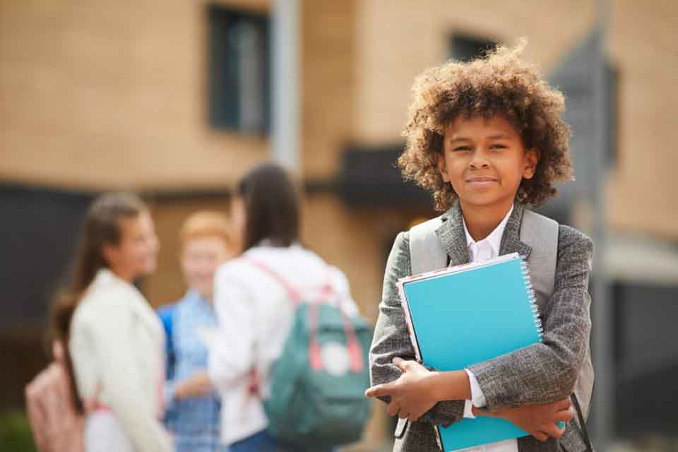 A student wearing a backpack and holding schoolbooks while standing in front of a school.