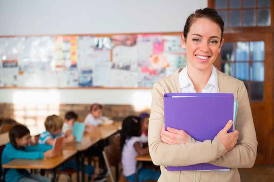 A teacher stands in an elementary school classroom holding a folder.
