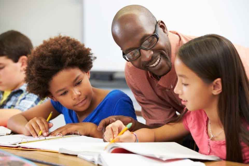 A smiling teacher leans forward to help two students with open notebooks working at a classroom table.