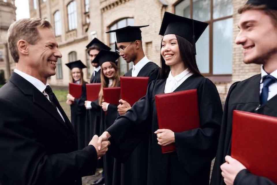 A man smiling and shaking hand with a student, who's dressed for a graduation ceremony