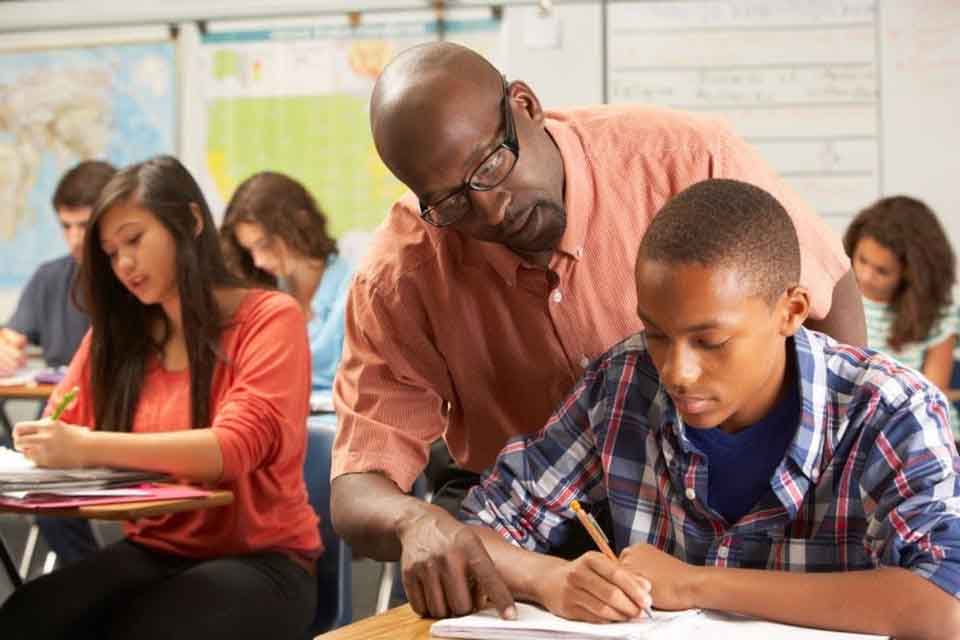 African American teacher in high school classroom looks over the shoulder of one of his students, an African American teenage boy, and points to his notebook.