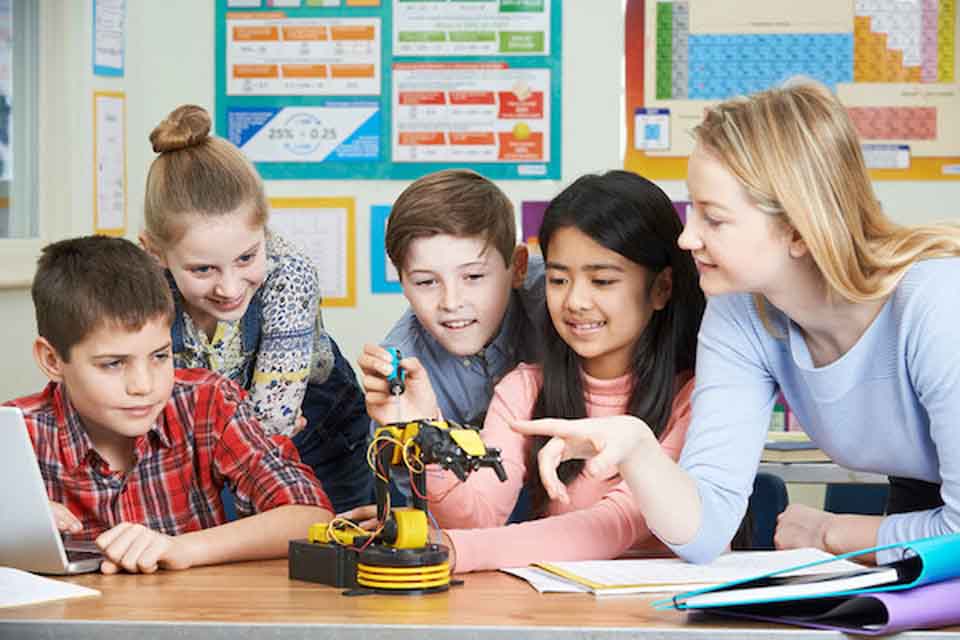 A woman pointing at a small robot. Four children are smiling and looking at the robot.