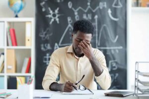 A tired teacher holds a hand to their forehead while sitting at a desk.