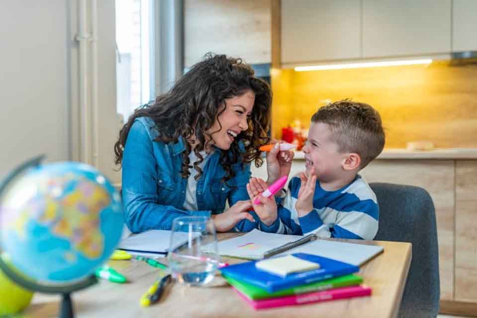 A casually dressed teacher laughs with a little boy at a table cluttered with books, notebooks, markers, and a globe.