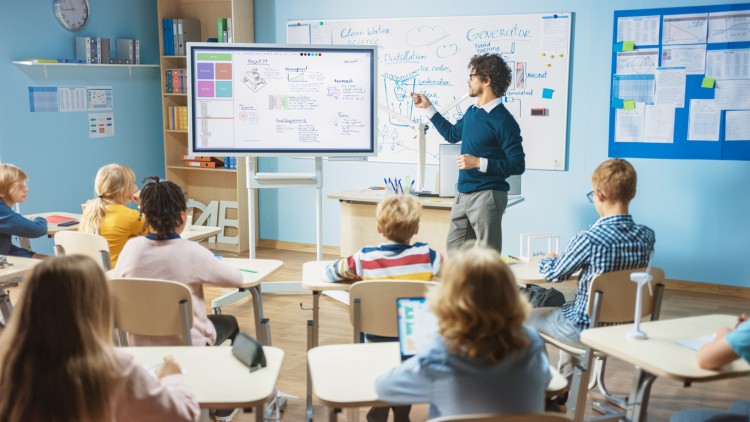 A teacher standing in front of a classroom points to a digital whiteboard.
