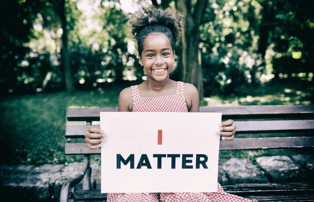 A smiling Black preteen girl sitting on a park bench holds a sign that reads "I matter."