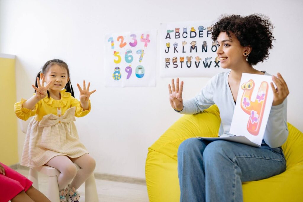 A smiling preschool teacher seated in front of several young children holds a card with an illustration of the number four in one hand and, with the other, shows four fingers.