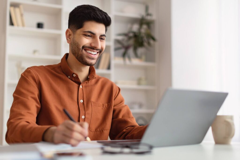 A man wearing a brown shirt sits at a desk, using a laptop computer and taking notes.