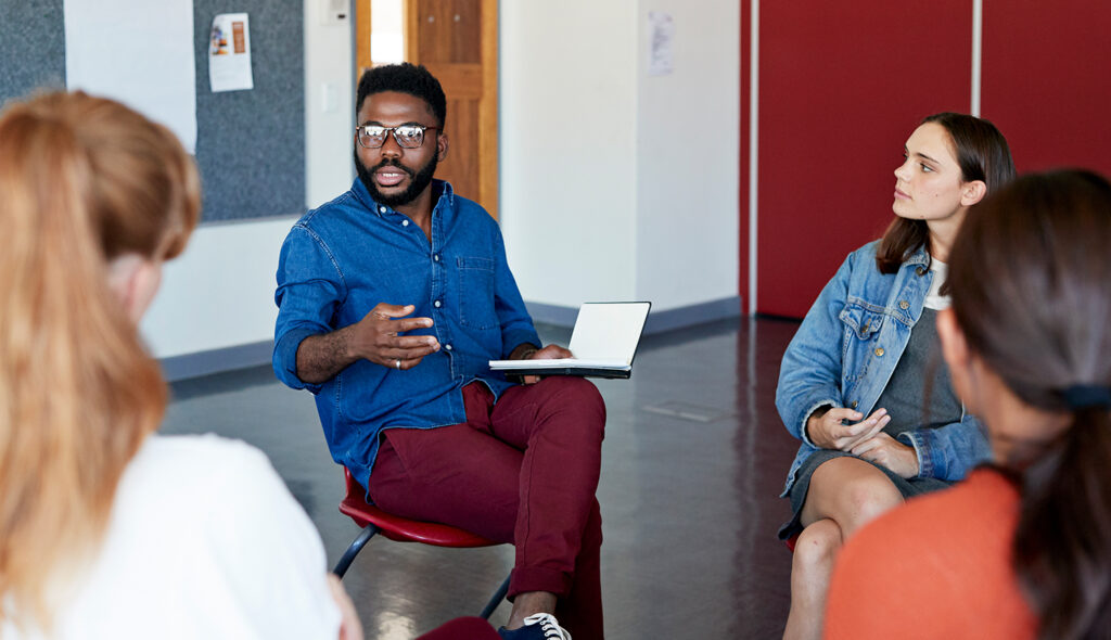 A professional educator holds an open notebook and facilitates a group discussion in a circle of adults.