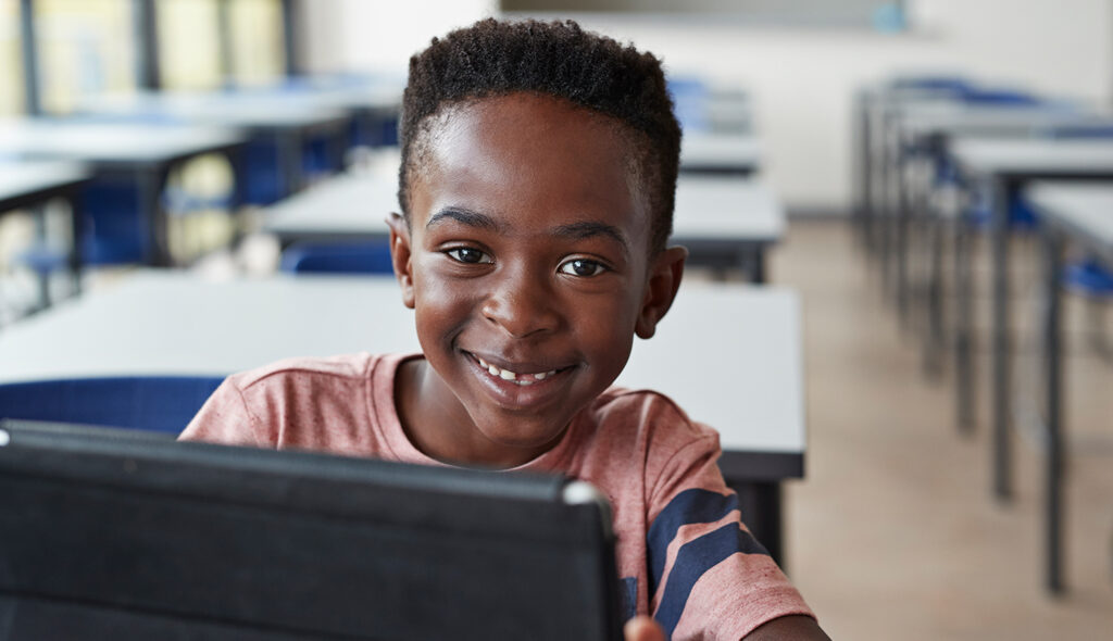 A child smiles at the viewer while working on a computer in a classroom.