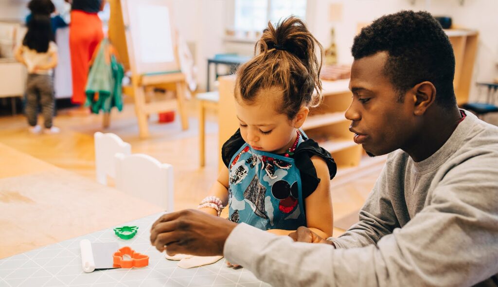 A teacher kneels down beside a preschool student who works with clay and various plastic molds.