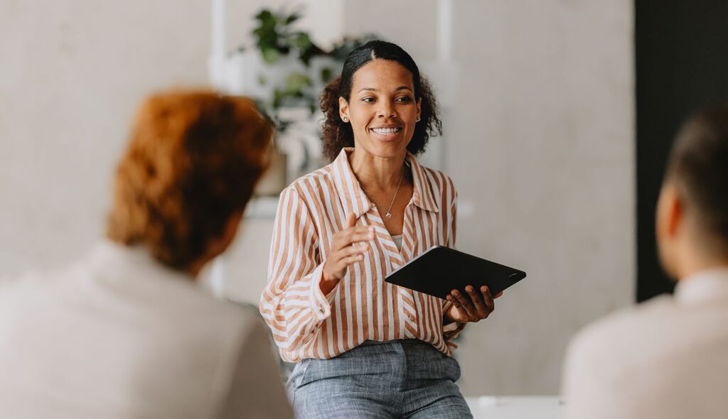 A woman holding a tablet presents to a group of colleagues.