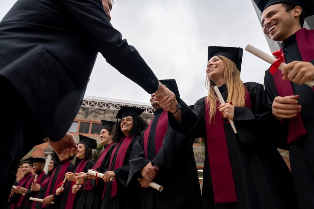 A school administrator shakes hands with smiling graduates in caps and gowns standing in a reception line.