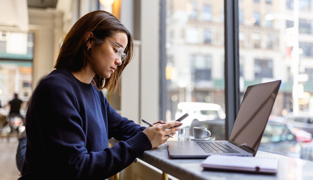 A graduate student wearing glasses sits in front of an open laptop and notebook while studying something on her phone.