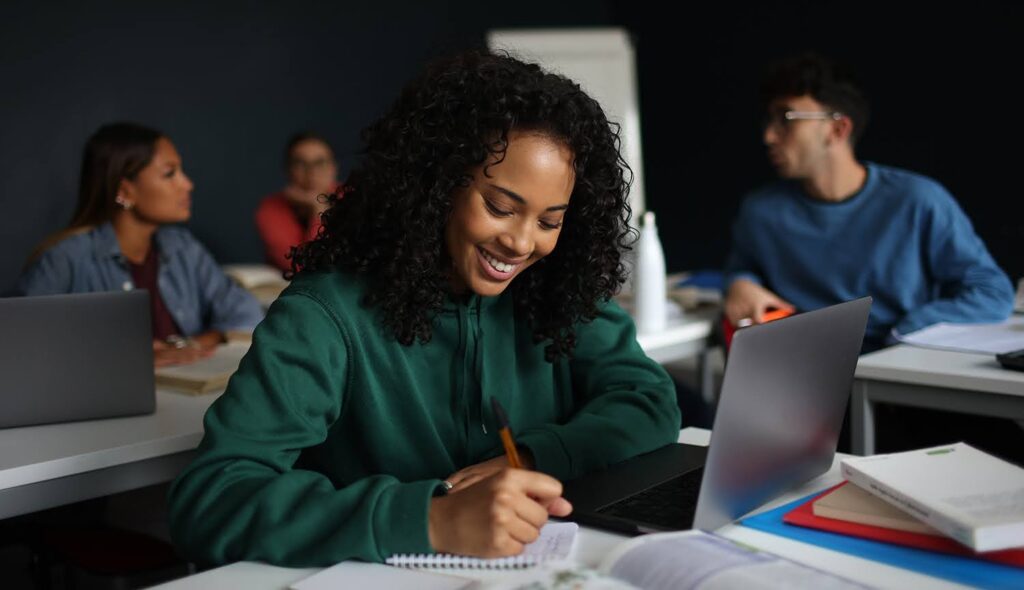 A student smiles as she writes in her notebook surrounded by other students in a classroom setting.