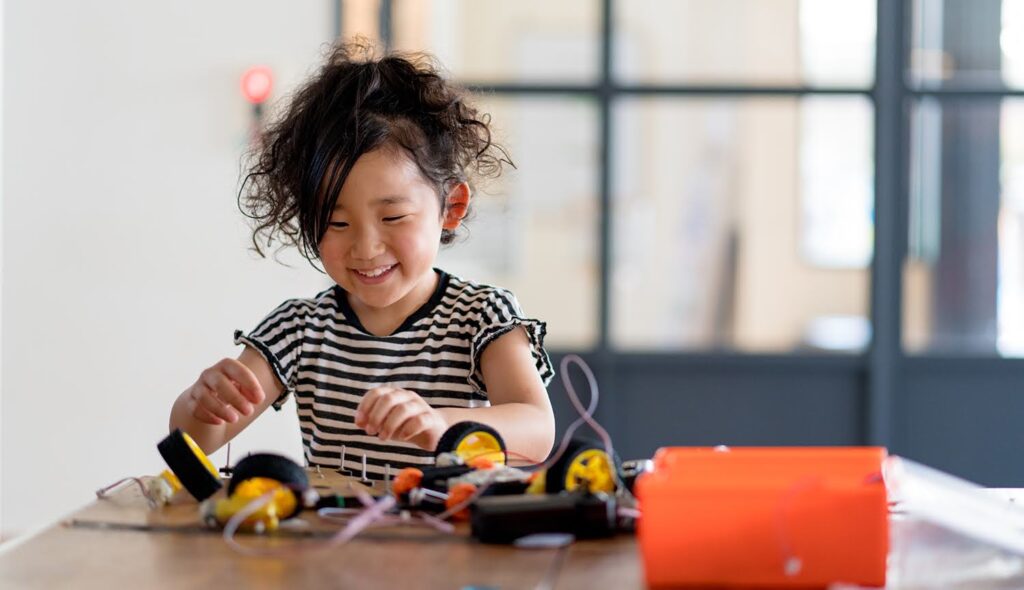 A young elementary school student smiles as she assembles the components of a project on her desk.