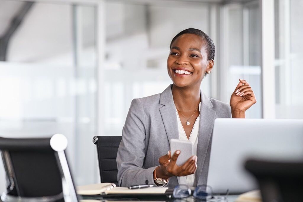 A woman in business attire sits at a conference table, looking up from the cell phone in her hand. Her laptop is open, and beside it are an open scheduler and pen.