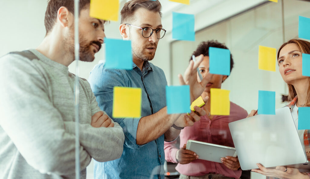 Four colleagues examine a transparent board filled with differently colored sticky notes.