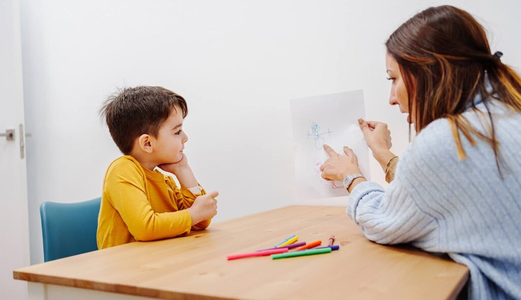 An educator points to a drawing on a piece of paper as she speaks to a young student.