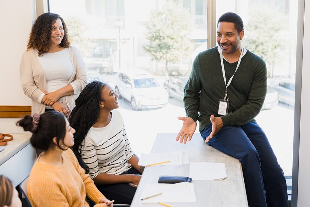 A smiling educator in a classroom leads an engaging discussion with an attentive group of students.