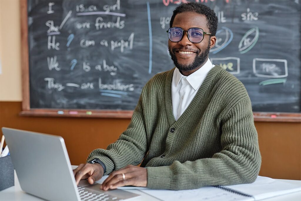 A smiling teacher sits at a desk with a laptop and papers; in the background, a blackboard features an English lesson.