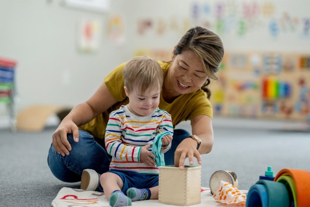 A teacher and young child sit on the floor in a colorful classroom, playing with a sensory toy box and engaging in hands-on learning activities.