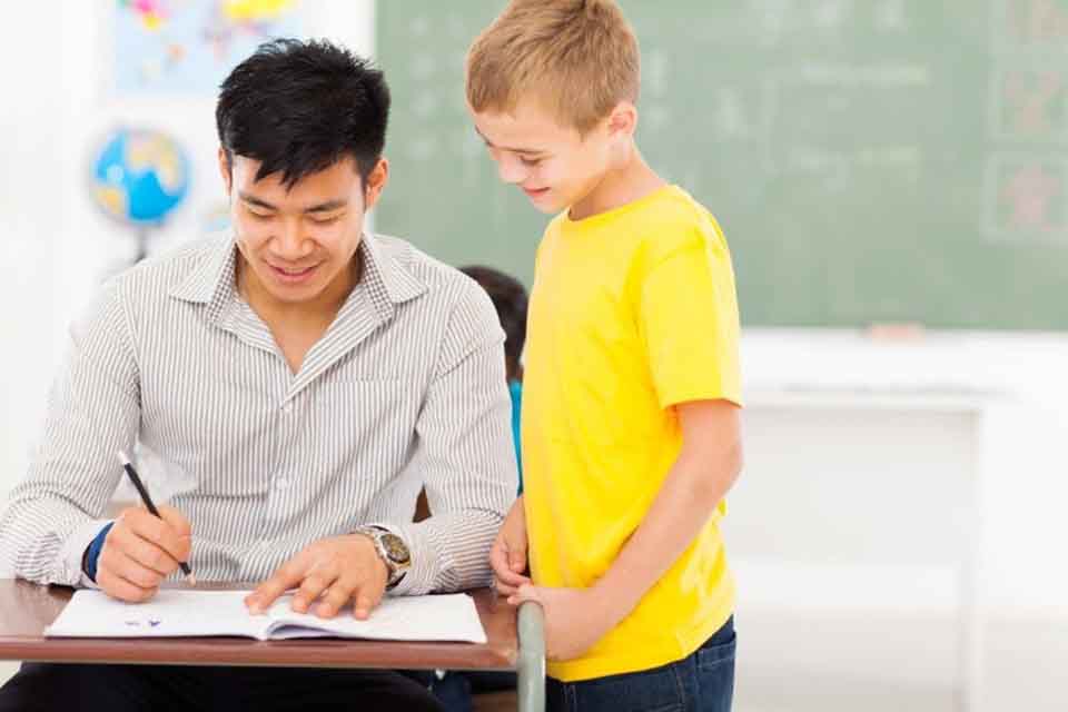 A smiling teacher sits at a school desk grading an elementary school student’s portfolio while the student looks on.