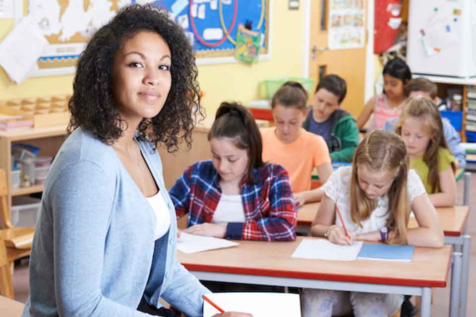 A teacher looking at the camera while the students write on a paper on their desks