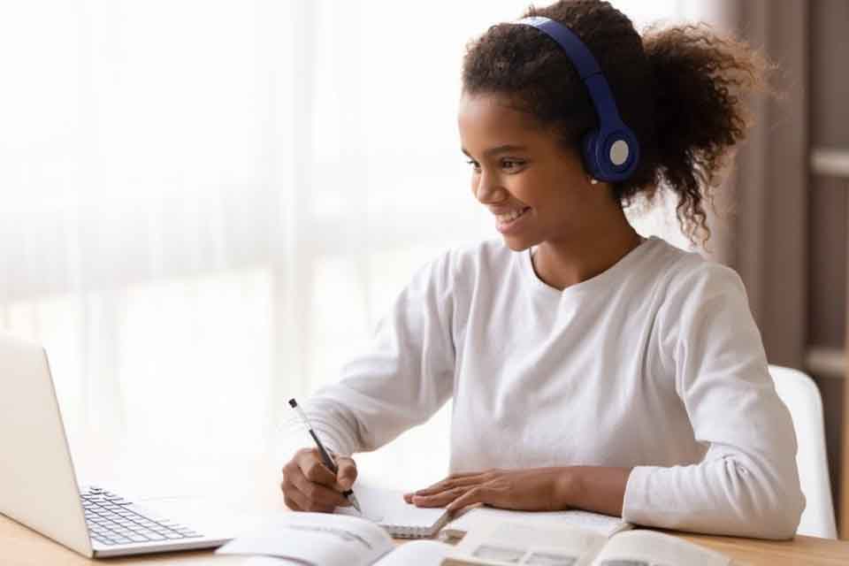 A smiling young Black student wearing headphones is taking notes while seated in front of her laptop computer.