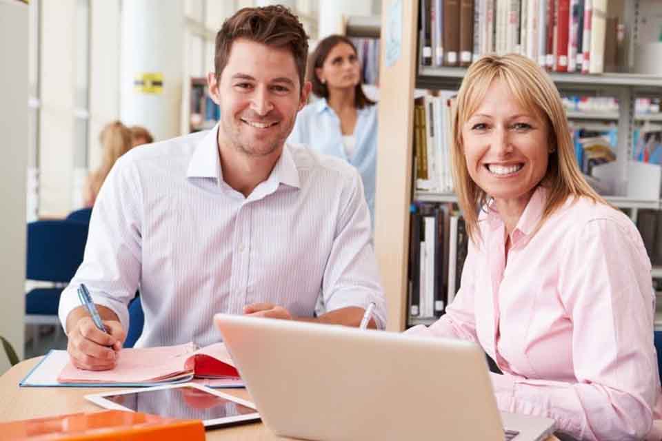 Two smiling teachers sit in the school library, one with a laptop, the other with a tablet and notebook.