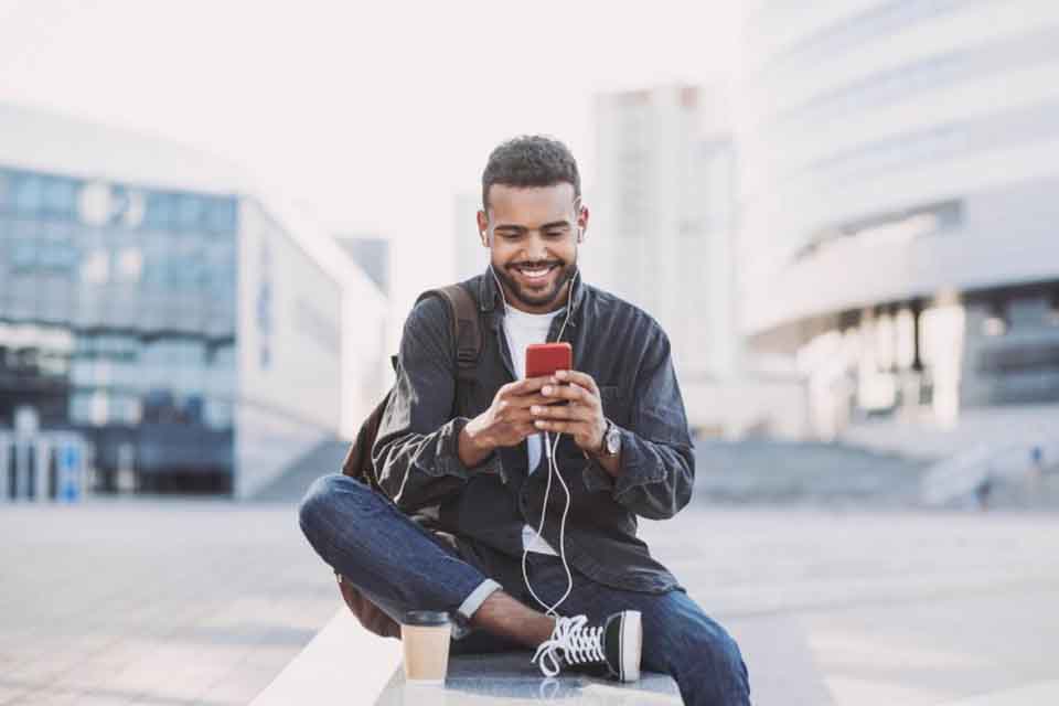A smiling person sits on a wall in an urban setting looking at a smartphone screen.