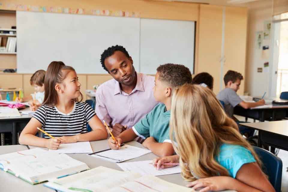 A teacher talks to three elementary school students who are working at a table in a diverse classroom setting.