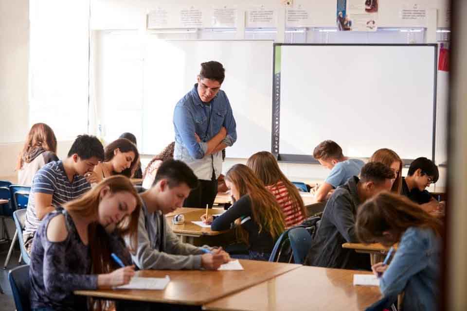 A high school stands among students working at their desks.