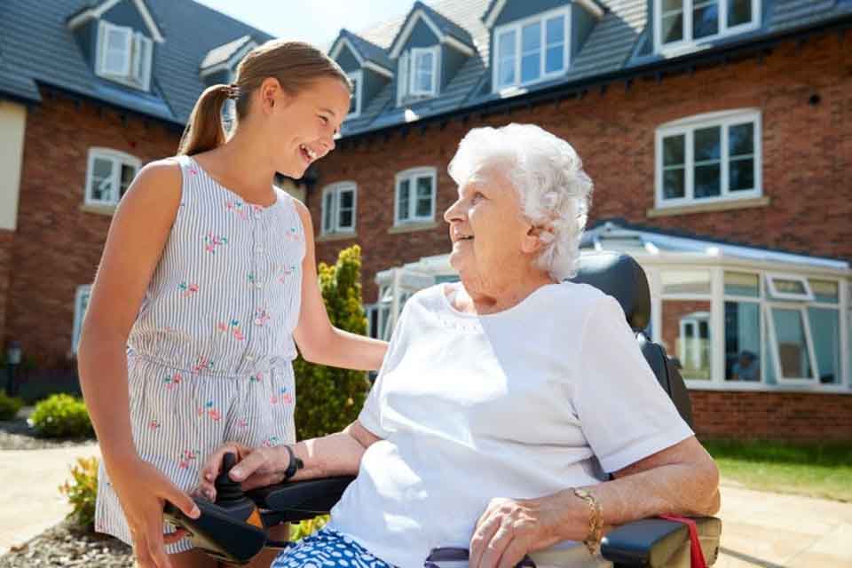 A young girl volunteers at a senior center and talks to an elderly woman outside.