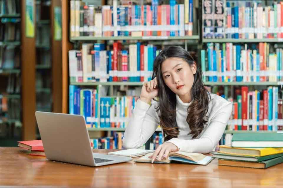 A woman sits at a library table with stacks of books and a laptop.