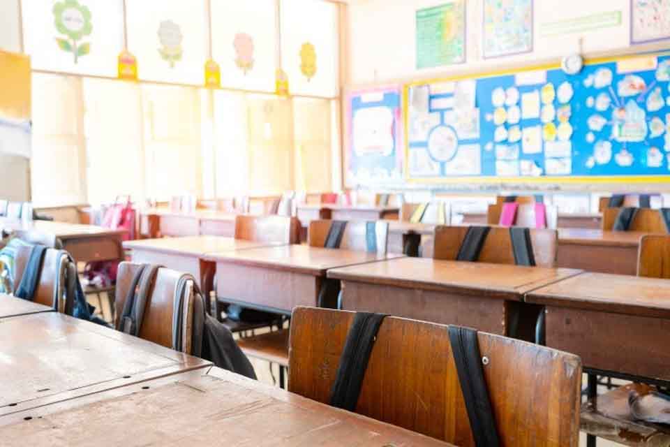 Backpacks hang on empty desks in a classroom.