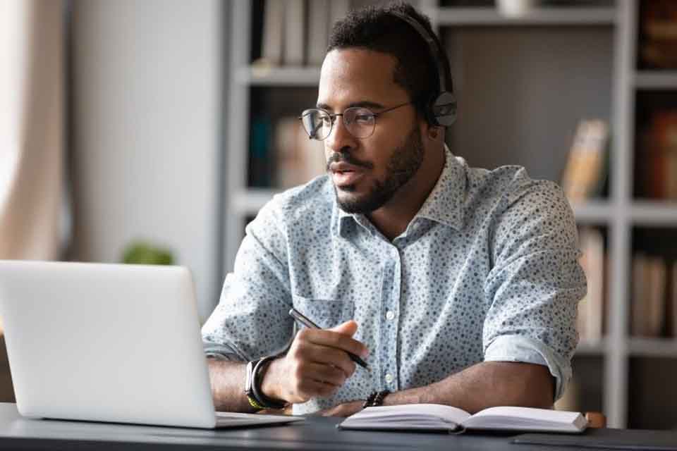 A teacher sits at a desk looking at a laptop and writing in a notebook.