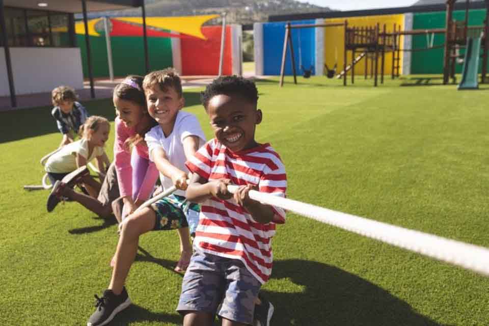 A group of smiling elementary school children play tug of war on the school playground in the summer.