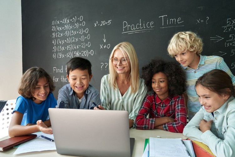 An elementary school teacher and a group of students look at a laptop.