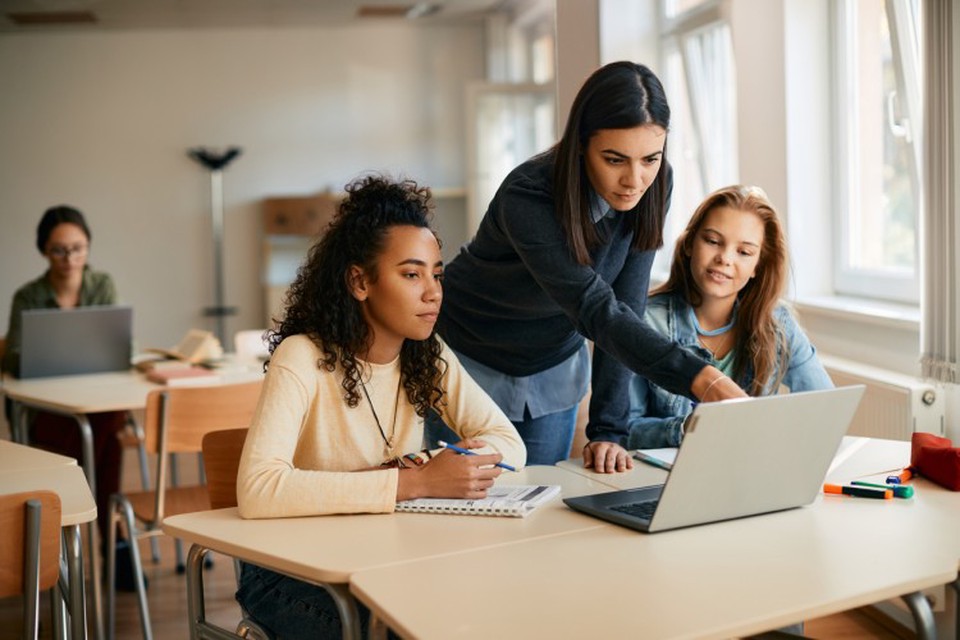A teacher stands between two students and points at a laptop.