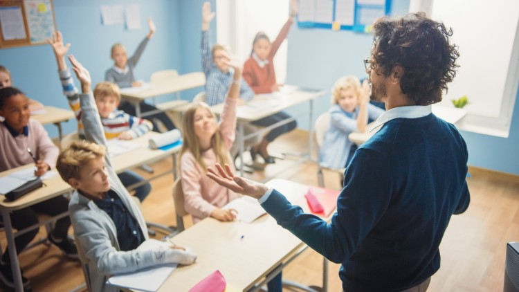 A middle school teacher stands in a classroom facing students raising their hands.