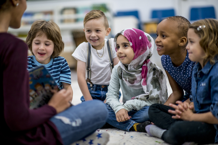 School children sit on the floor and listen to their teacher read a storybook.