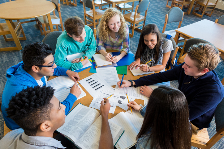 High school students gathered around a table listen to a teacher.