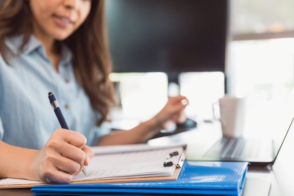 Closeup of an educator concentrating while working at a laptop and taking notes on a notepad.