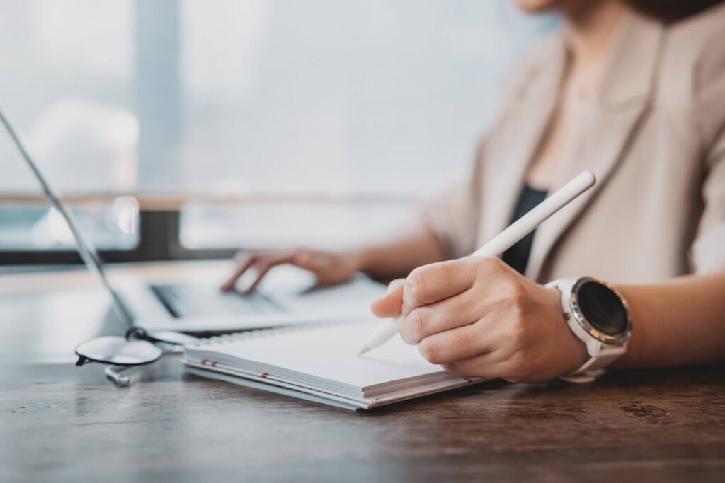 A professional in business attire takes notes while working at a laptop.