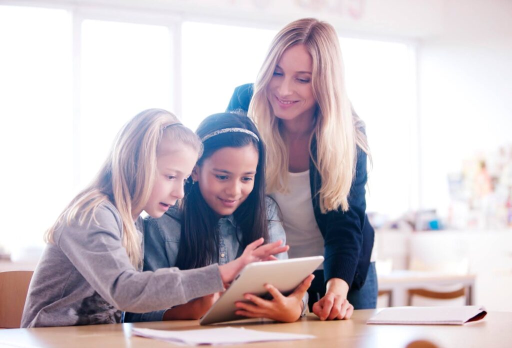 A teacher watches while two young students use a tablet computer.