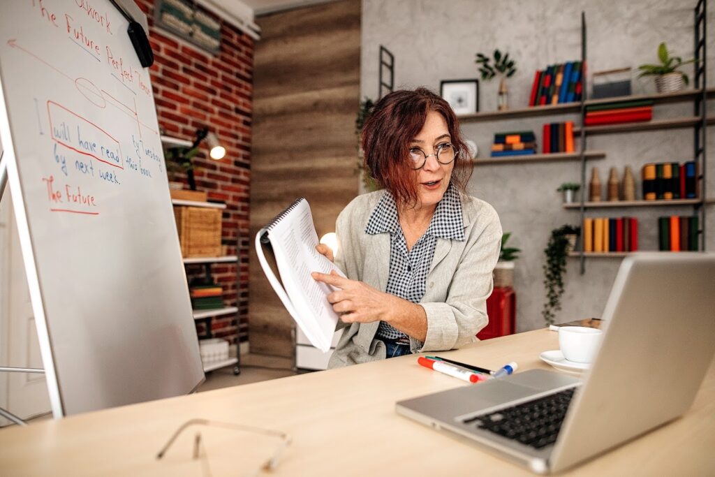 An English language teacher delivers a lesson remotely via teleconferencing; a sentence diagram identifying verb tenses is visible behind her.